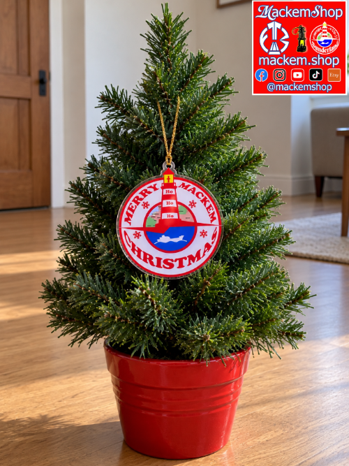 A Christmas tree ornament hanging on a small artificial Christmas tree in a red pot, placed on a wooden floor in a living room. The ornament features a lighthouse, blue water, and the text 'MERRY MACKEM CHRISTMAS'. A MackemShop Logo is visible in top right corner.
