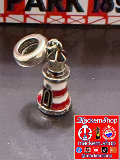 A silver-toned Roker lighthouse charm with red and white enamel stripes, shown on a dark surface with a red and white sign blurred in the background. A 'MackemShop' logo in right downside corner.