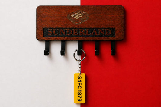 A wooden key holder with black hooks, engraved with 'SUNDERLAND' and a crest, holding a yellow luggage tag keychain that reads 'S4FC1879'. The background is split vertically with white on the left and a vibrant red on the right.
