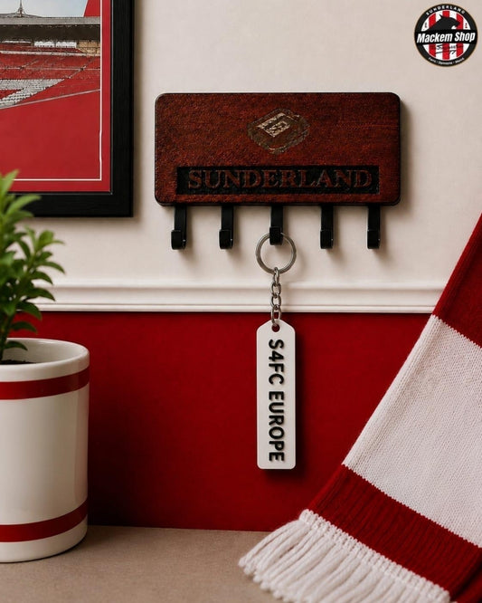 A wooden Sunderland AFC Europe key holder mounted on a wall, featuring a hanging white tag that reads 'S4FC EUROPE'. The scene includes a red and white striped scarf, a potted plant, and a framed picture, with a Macken Shop logo in the top right corner.
