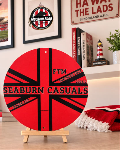 A circular red and black Seaburn Casuals FTM Metal sign on a small wooden easel stand, displayed on a tabletop with Sunderland-themed decor in the background.