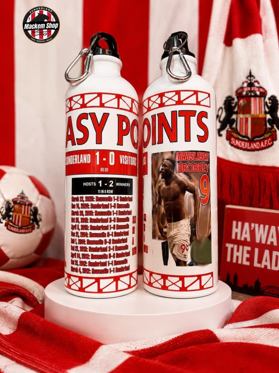 Two white water bottles with red and black Sunderland AFC football graphics, displayed on a white pedestal with red and white team-themed fabric and a soccer ball in the background.