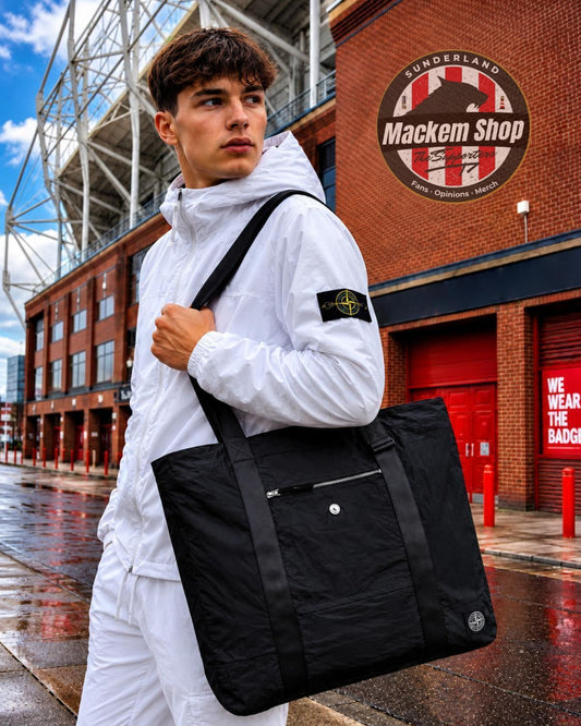 A man in a white Stone Island tracksuit carries a black Stone Island tote bag with the brand's compass patch visible on his sleeve, set against a wet street scene in front of a brick building with a 'Mackem Shop' sign.