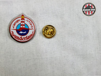 Two pins on a textured gray background, one with a lighthouse design and text, the other gold.