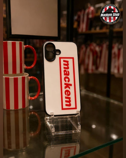 A white phone case with a red 'mackem' logo displayed on a clear stand on a glass shelf, with red and white striped mugs in the background.