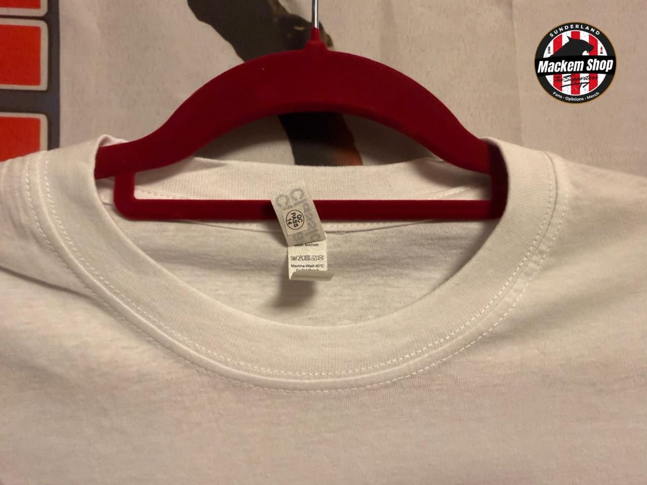 White t-shirt on a red hanger with a visible brand tag, against a neutral background.