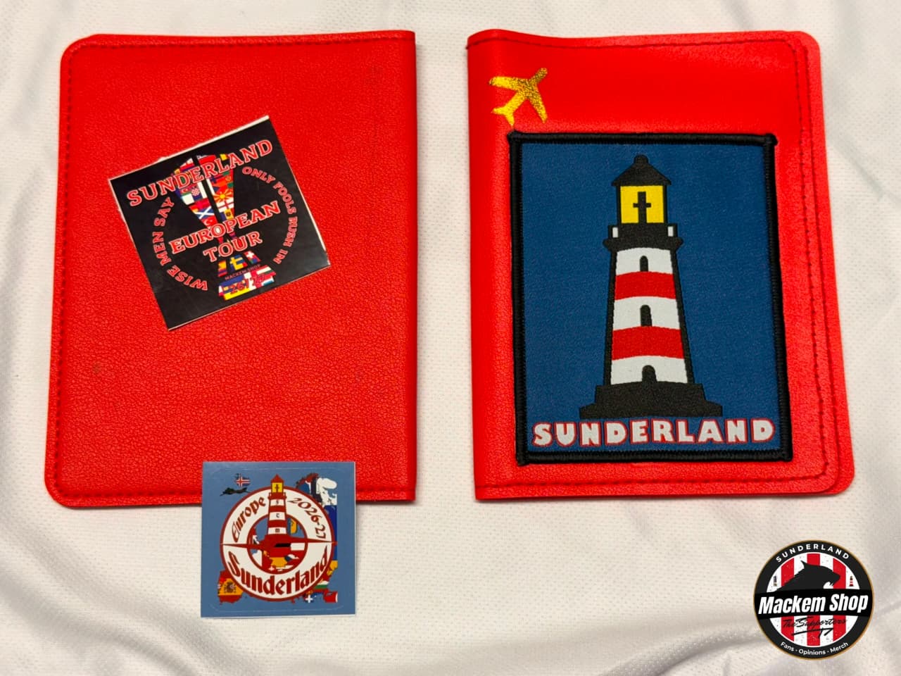 Two red fabric Passport covers displayed on a white fabric background, each featuring a Sunderland-themed design. One shows a black square patch with red and white Sunderland text and imagery, and the other shows a blue square patch with a red-and-white lighthouse and the word 'SUNDERLAND.