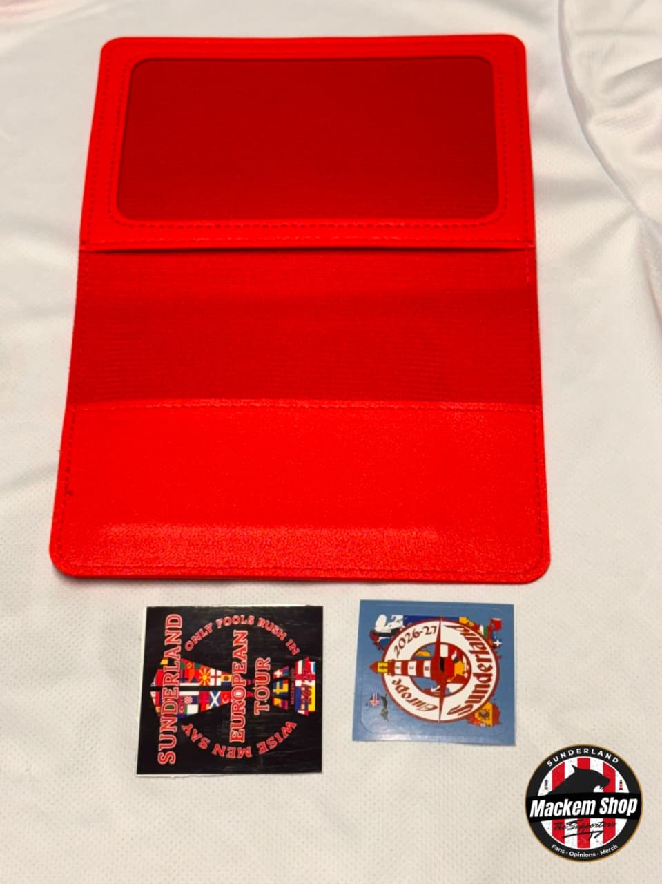 A bright red fabric passport holder laid flat on a white fabric background, with two small, printed patches or stickers placed below it. A circular 'Mackem Shop' watermark appears in the bottom right corner.