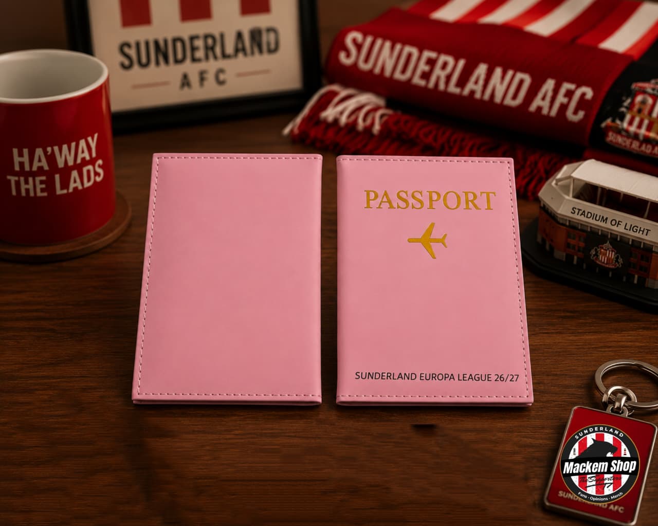 A pink faux leather passport cover set shown on a wooden table, with one cover closed and one open to display the interior. The open cover features gold foil text reading 'PASSPORT' and 'SUNDERLAND EUROPA LEAGUE 26/27' with a small airplane icon.