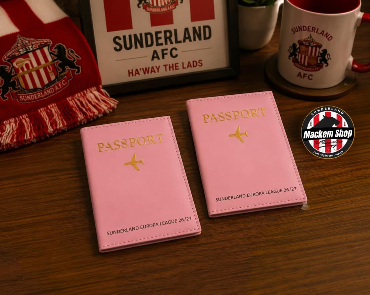 Two pink passport cover sleeves with gold foil lettering and airplane icons, displayed on a wooden table alongside Sunderland AFC themed merchandise including a scarf, framed sign, mug, and a MackemShop logo badge.