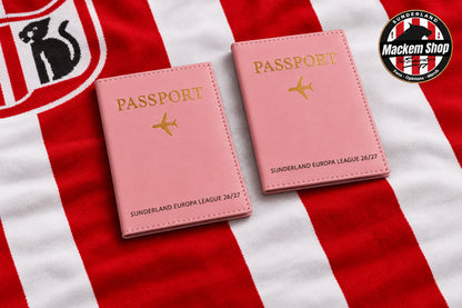 Two pink passport cover sleeves with gold foil lettering and airplane icons, displayed on a wooden table alongside Sunderland AFC themed merchandise including a scarf, framed sign, mug, and a MackemShop logo badge.