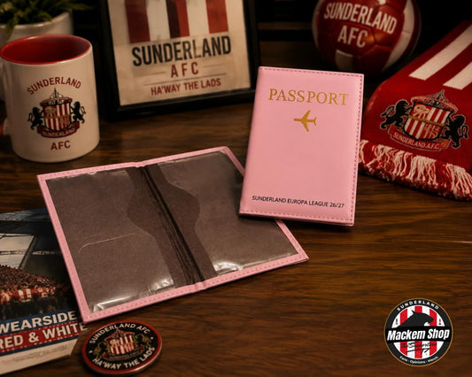 A pink faux leather passport holder set displayed on a wooden table, featuring a closed pink passport cover with gold lettering and an open matching pink sleeve with clear ID windows. The scene is styled with Sunderland AFC themed props including a mug, scarf, ball, and framed sign.