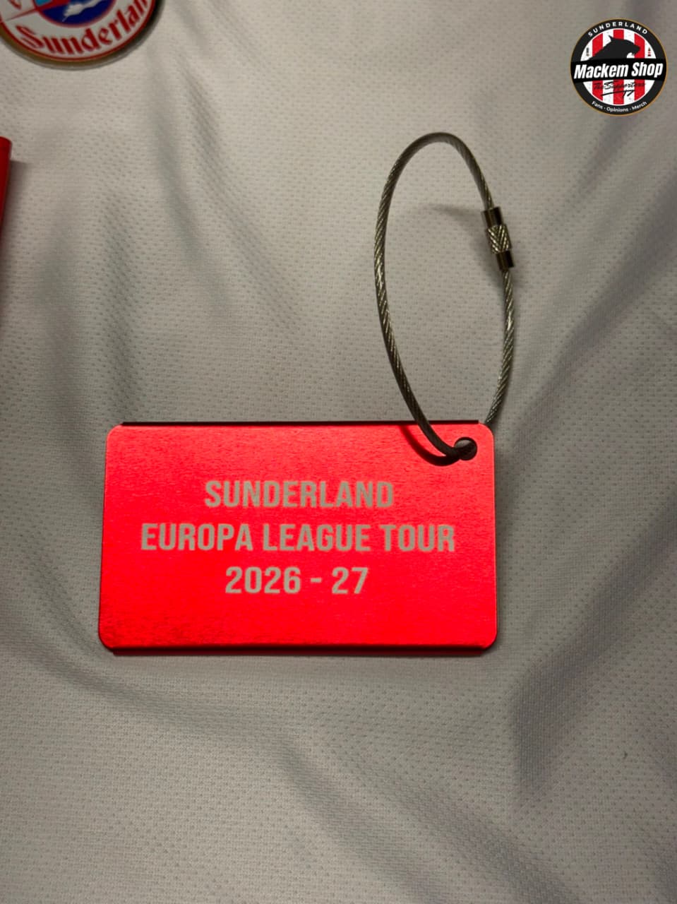 A red rectangular tag with a metal cable lock, printed with 'SUNDERLAND EUROPA LEAGUE TOUR 2026 - 27' in white text, placed on a light gray textured surface.