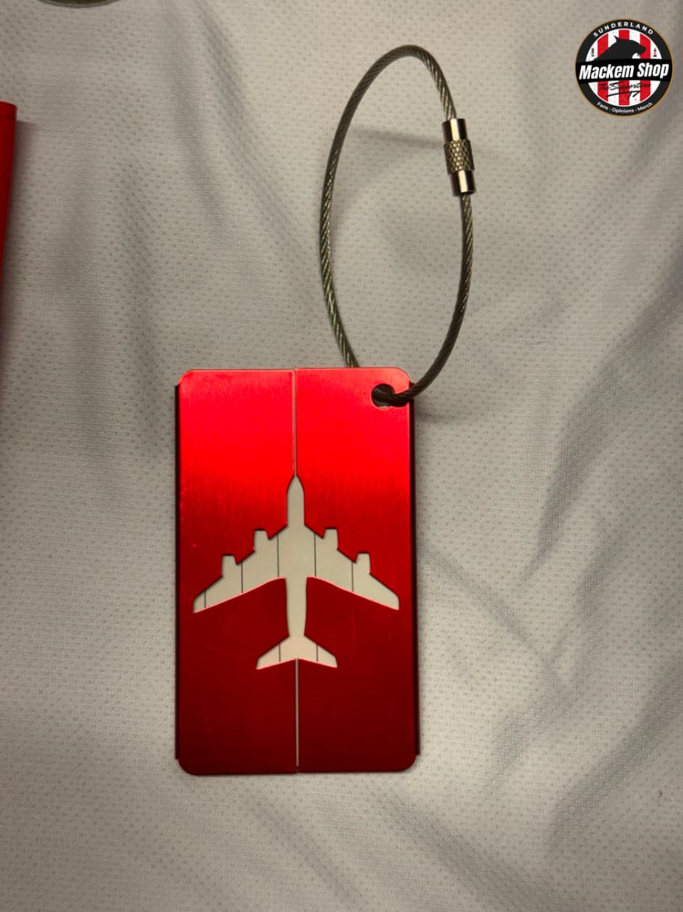 A red rectangular luggage tag with a white airplane silhouette, attached to a silver cable lock, displayed on a white textured fabric background.
