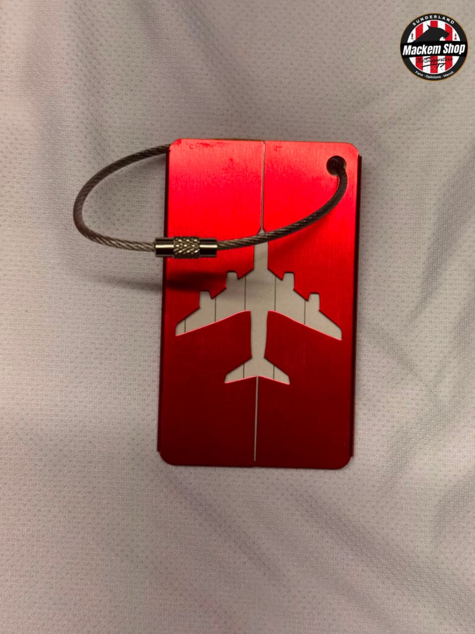 A red luggage tag with a white airplane silhouette in the center, attached to a dark metal cable lock, displayed on a textured white fabric background. A circular 'Mackem Shop' logo is visible in the top right corner.