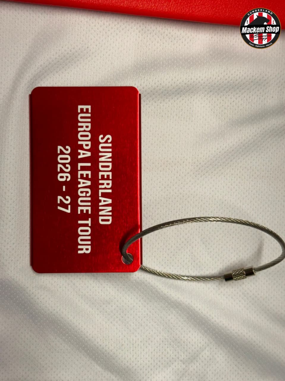 A red metal tag with white text reading 'SUNDERLAND EUROPA LEAGUE TOUR 2026 - 27' attached to a silver wire loop, placed on a white textured fabric background. A circular 'Mackem Shop' logo is visible in the top-right corner.