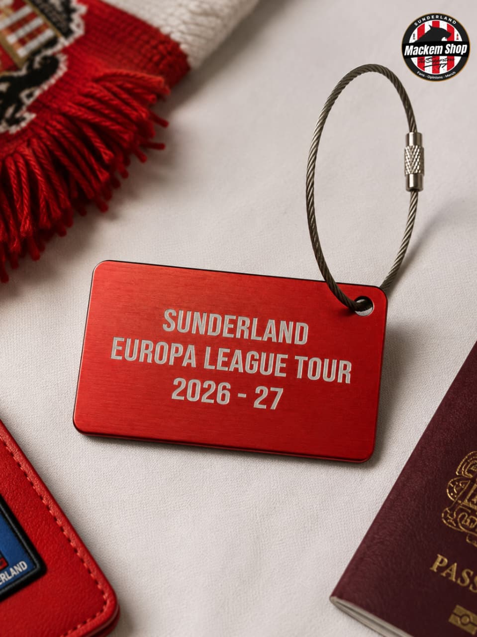 A red rectangular luggage tag with a silver wire loop, printed with 'SUNDERLAND EUROPA LEAGUE TOUR 2026 - 27' in white text, styled on a light fabric surface with a red scarf, passport, and wallet nearby.