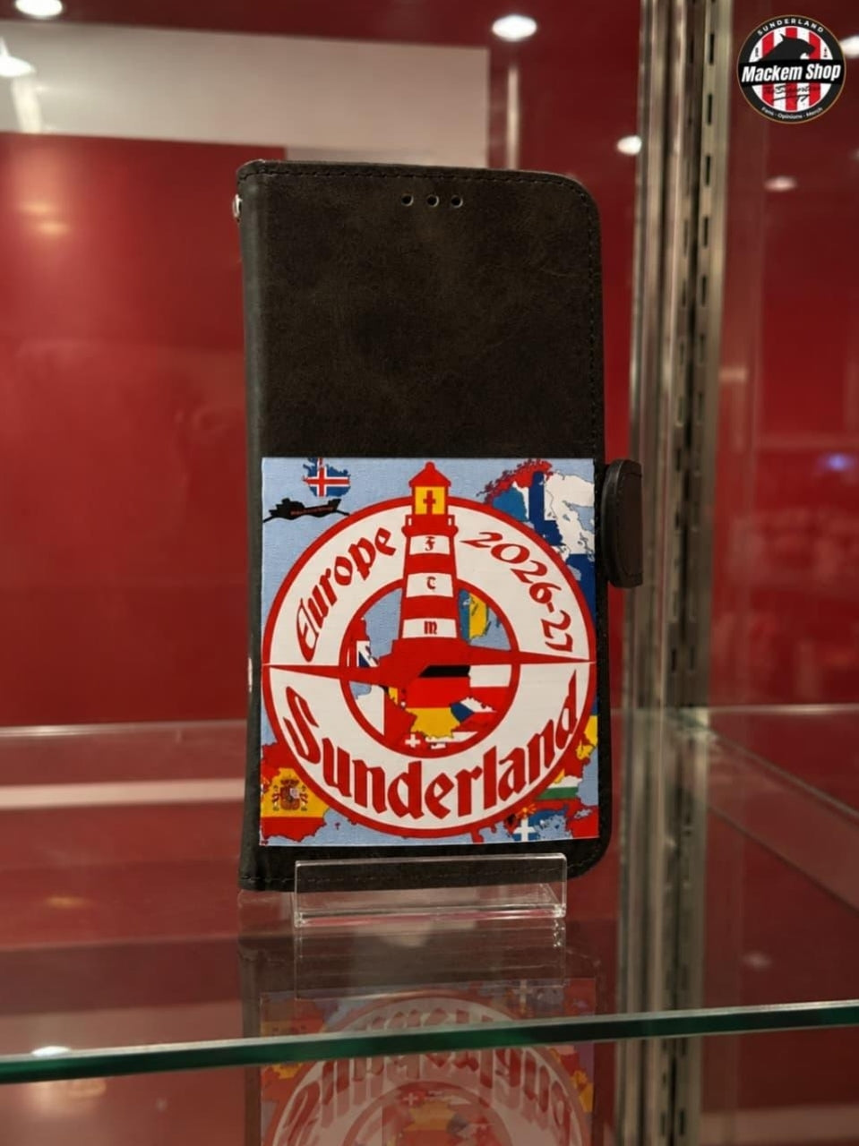 A black folio-style phone case with a colorful printed graphic on the Front, displayed on a glass stand inside a glass case. The graphic features a red and white lighthouse design with the text 'Europe 2026' and 'Sunderland'.