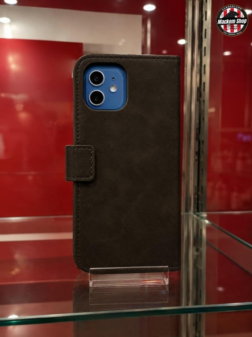 A Black faux-leather folio iPhone case with a magnetic tab closure, shown on a clear acrylic stand inside a glass display case with a red background.