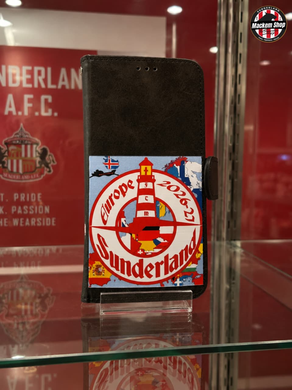 A black iPhone 12 case with a large Sunderland AFC graphic insert, displayed on a glass stand inside a red shop setting.