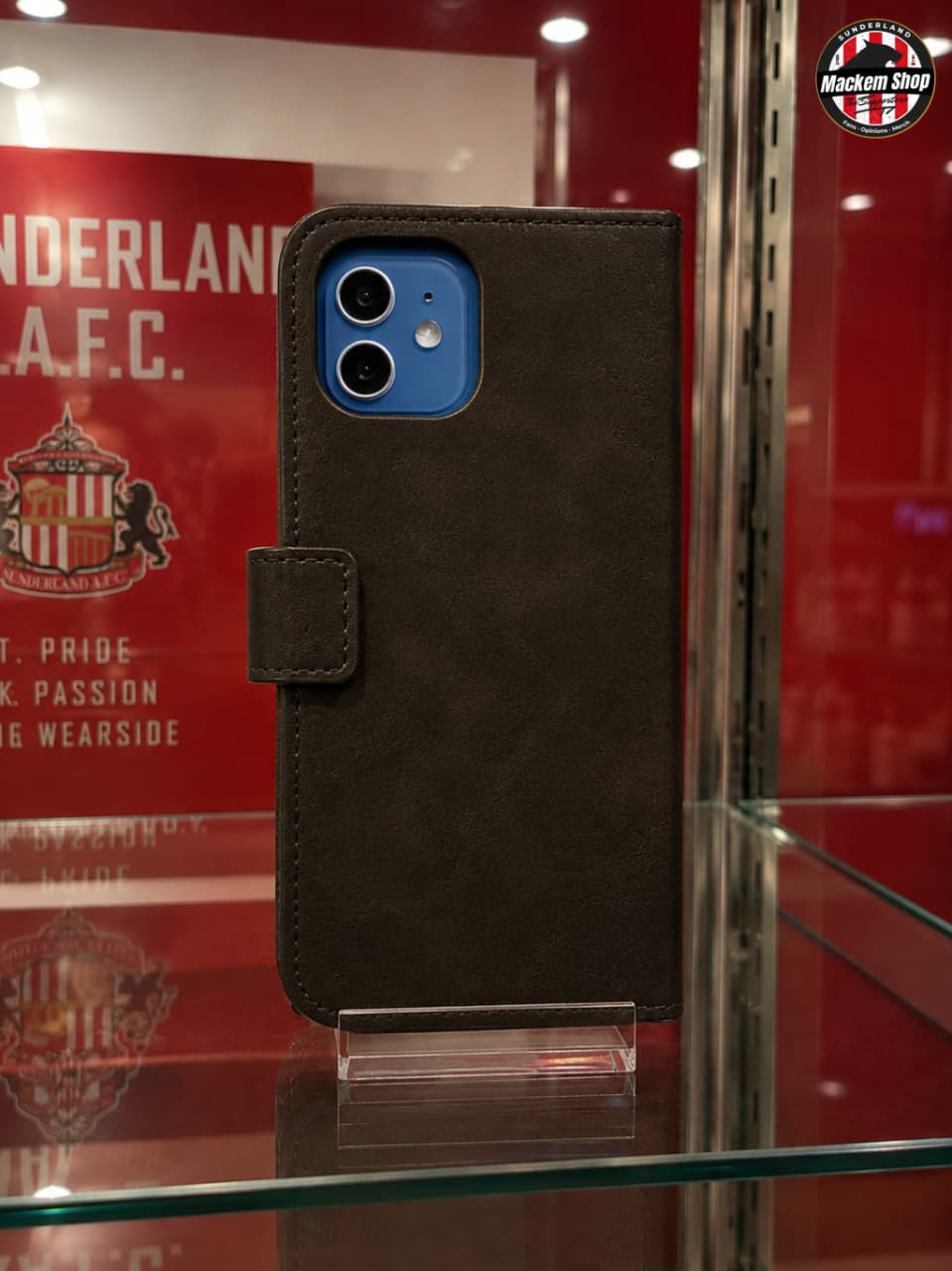 A black iPhone 12 case with a magnetic tab closure, shown on a clear glass display stand inside a glass case.