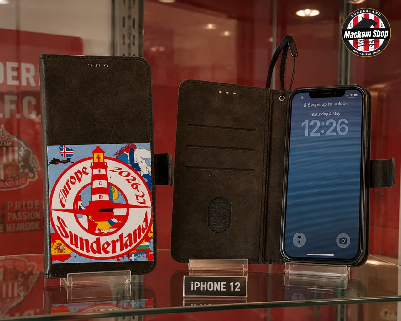 A black flip phone case shown open on a glass display case, with an iPhone 12 inserted. The left side of the case features a colorful printed graphic of a lighthouse and the text 'Sunderland Europe 26-27'.