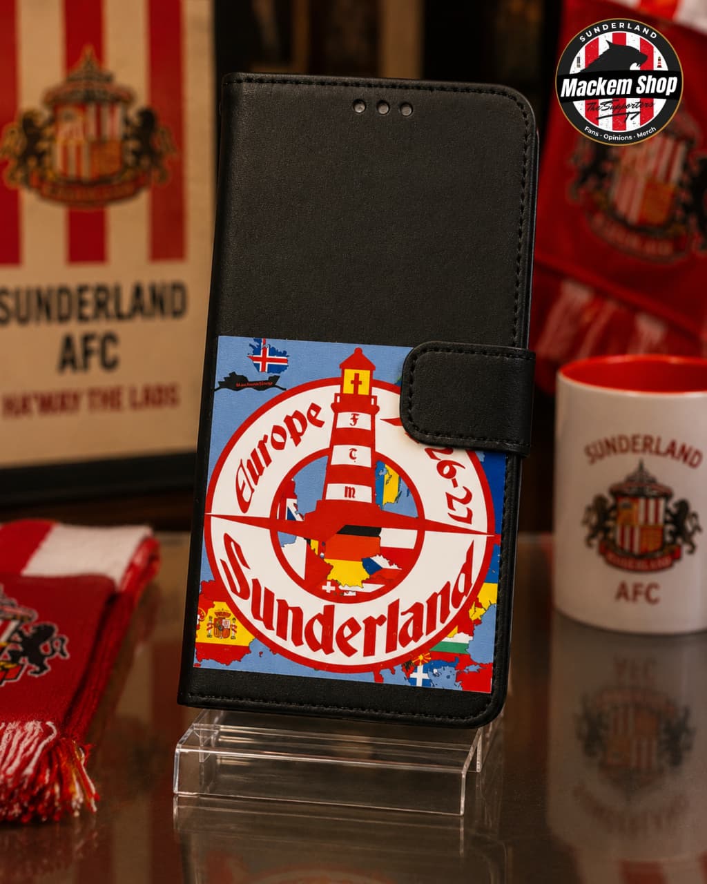 Black faux-leather wallet phone case featuring a colorful Sunderland AFC crest graphic, displayed on a clear acrylic stand with Sunderland-themed mugs and scarves in the background.