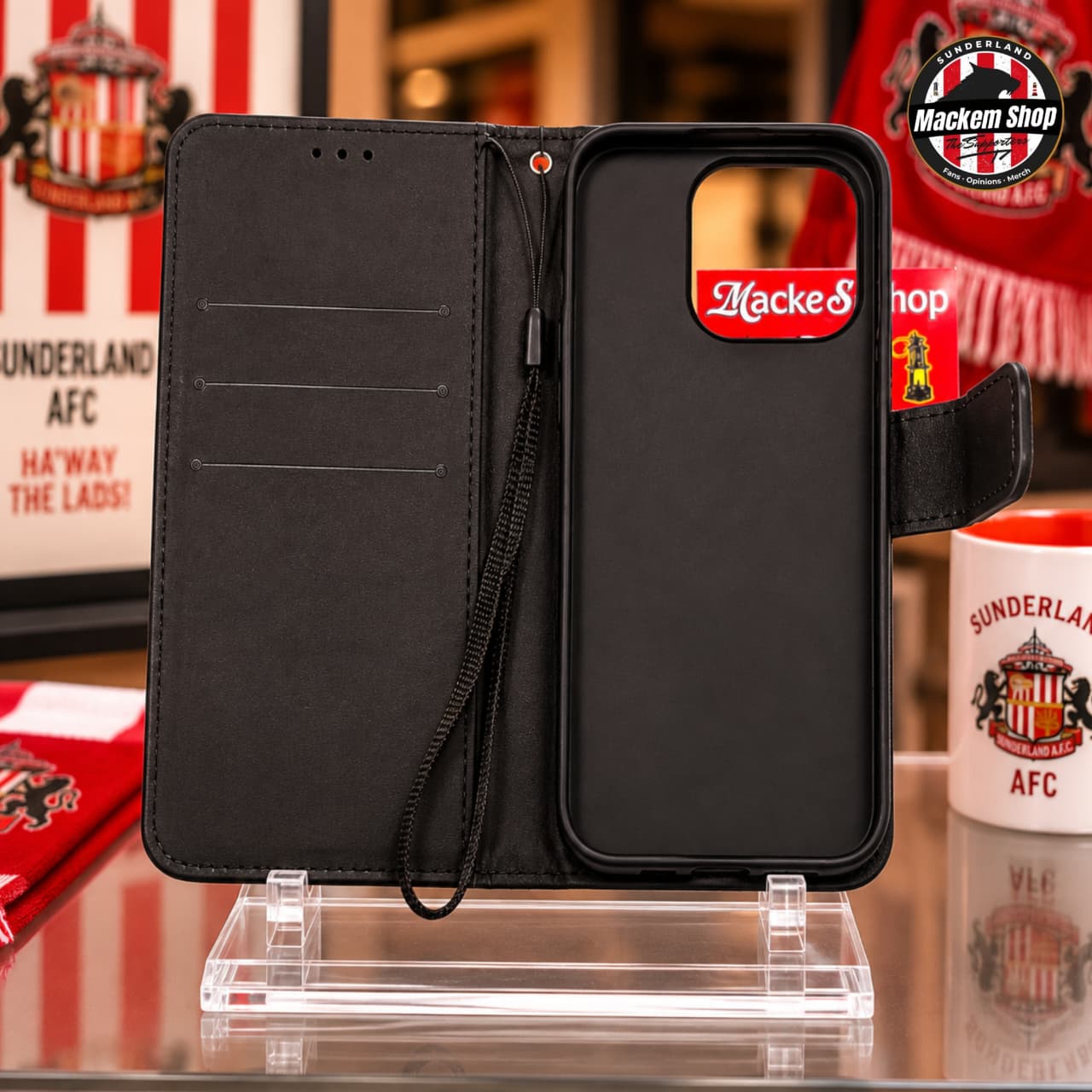 Black folio iphone case with a wrist strap, shown open on a clear display stand. The case has multiple card slots on the left panel and a camera cutout on the right panel, with a red and white 'Mackem shop' sticker on the back.