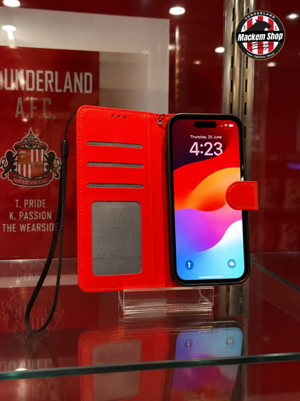 A red flip iPhone case with a black wrist strap, shown open with a smartphone inserted on a glass display stand. The case has card slots, a transparent ID window, and a small red tab on the back.