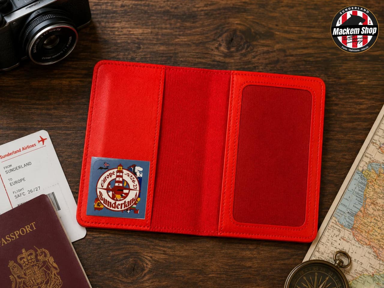 A red leather passport cover laid open on a dark wooden tabletop, with a small round London-themed patch on the left panel and a clear ID window on the right. A passport, boarding pass, camera, map, and compass are partially visible around it, and a 'Mackem Shop' logo appears in the top-right corner.