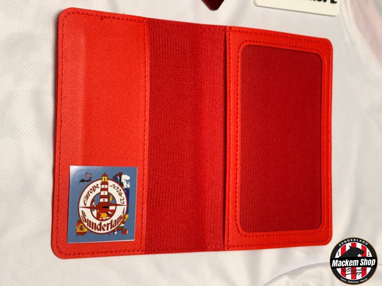A red leather passport cover shown open on a white textured surface, featuring a lighthouse patch on the left side and a clear ID window on the right.