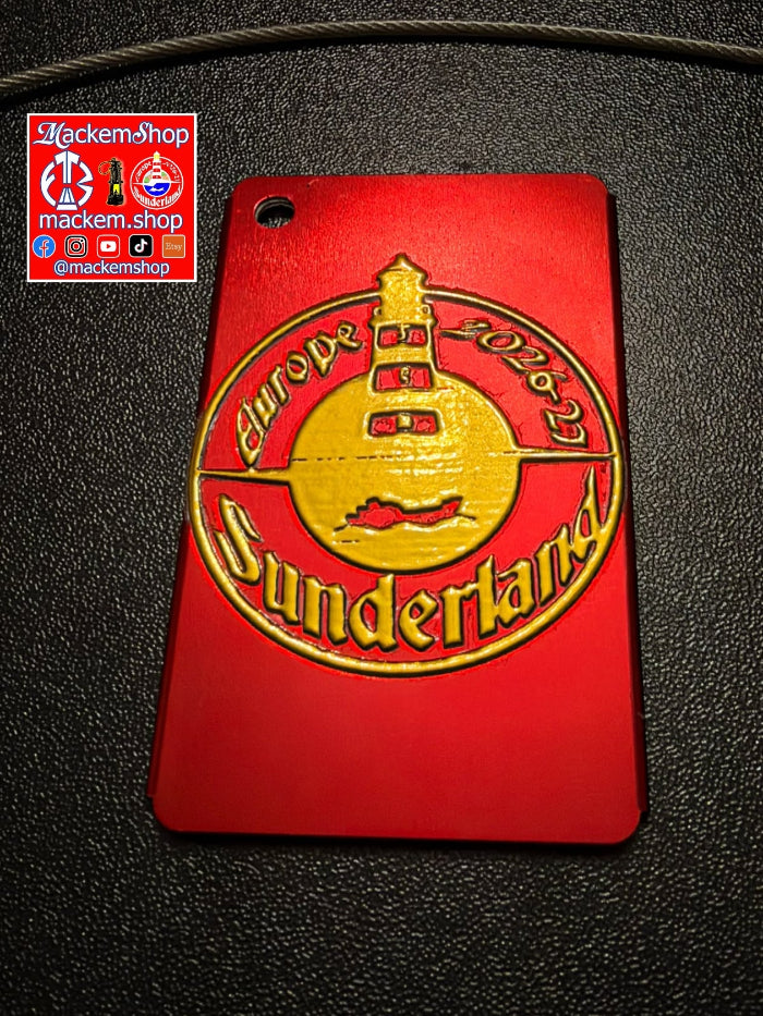 A red rectangular Luggage tag with a gold embossed design featuring a lighthouse and the words 'Europe 2026 Sunderland', photographed on a dark textured surface. A 'mackemshop' logo sticker is visible in the top-left corner.