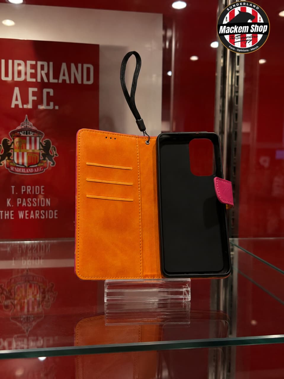 An orange folio-style phone case with a black interior and a wrist strap, displayed on a glass stand in a retail setting. A Sunderland A.