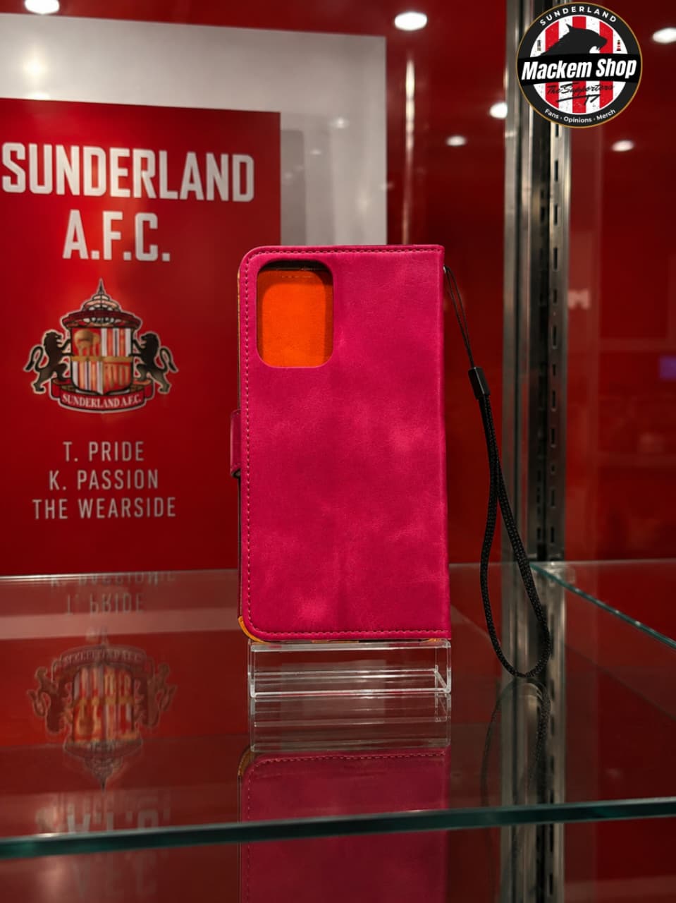 A bright pink suede-textured phone wallet case with a black wrist strap, displayed on a clear acrylic stand inside a glass case. The background features a red Sunderland A.