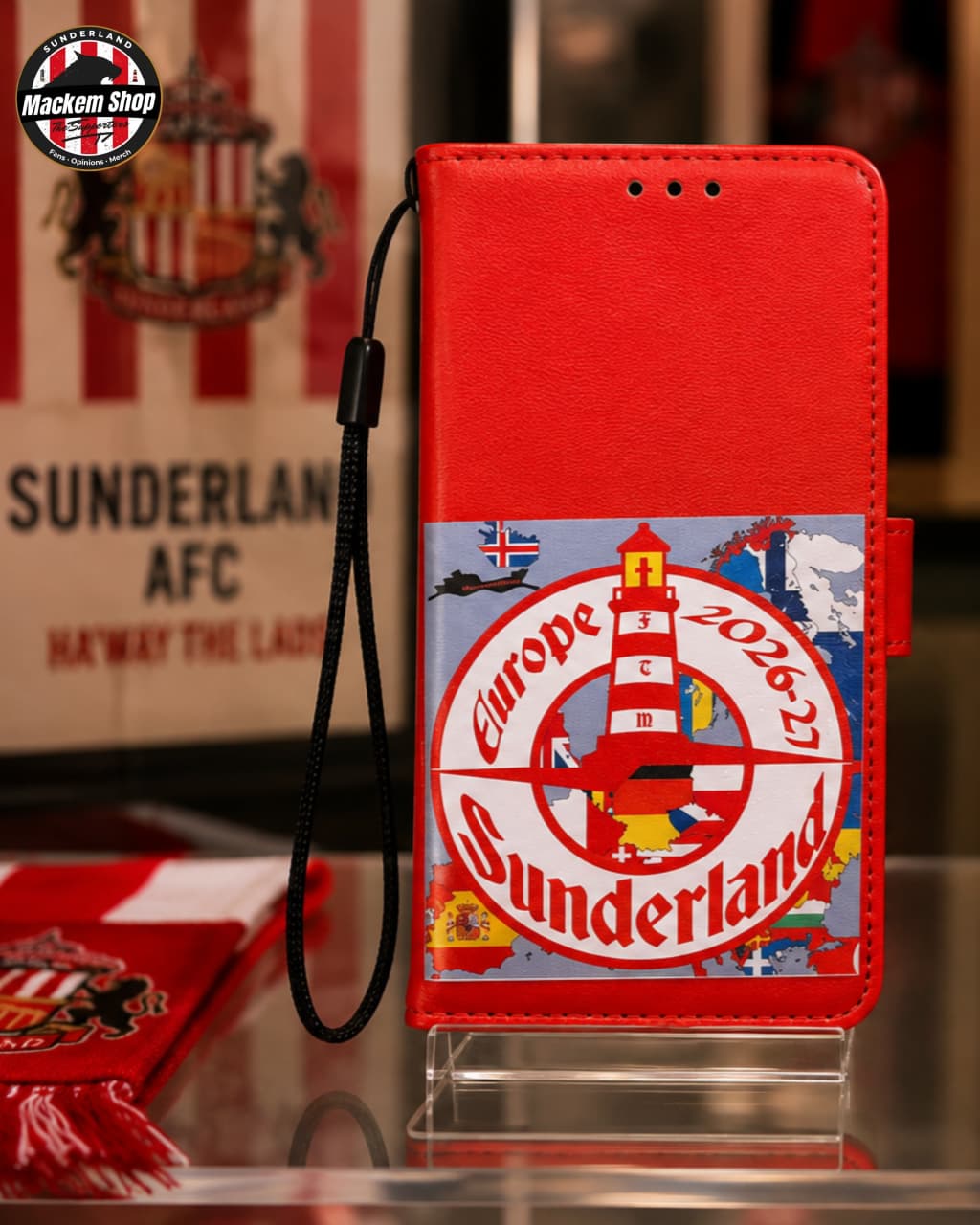 A red iPhone case with a Sunderland AFC theme, featuring a lighthouse graphic and 'Europe 2026' text, displayed on a clear stand with a Sunderland AFC scarf in the background.