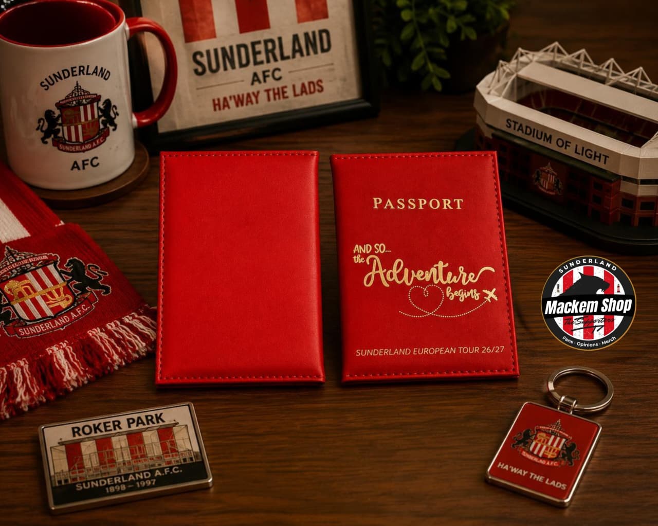 A red faux-leather passport holder and matching envelope set displayed on a wooden table, surrounded by Sunderland AFC themed merchandise including a scarf, mug, stadium model, and badges.