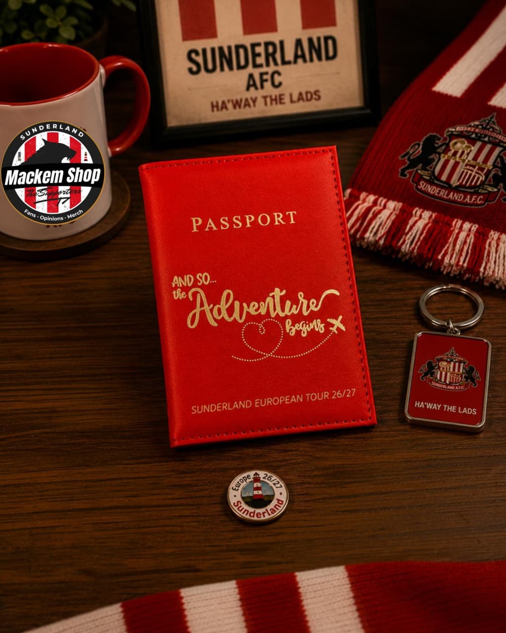 A red Sunderland AFC passport-style holder or mini passport case with gold lettering, displayed on a wooden table alongside a red-and-white striped scarf, a mug, a keychain, and a round badge. A Sunderland AFC sign is visible in the background.
