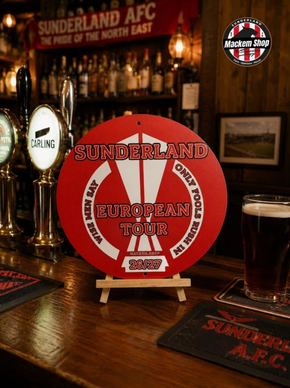 A round red and white Sunderland AFC European Tour 2027 Metal sign displayed on a wooden bar stool in a pub setting, with beer taps, a pint of dark beer, and Sunderland AFC coasters visible.