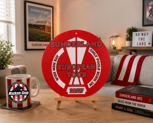 A round red and white Sunderland European Tour 2024/25 metal sign displayed on a small wooden easel in a cozy living room setting.