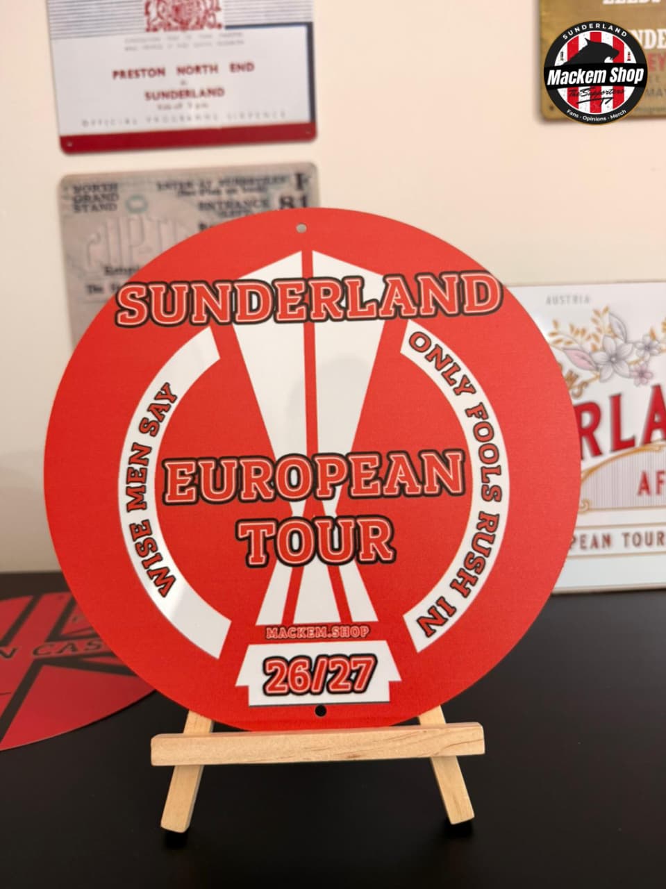 Red and white circular metal sign with 'Sunderland European Tour' text on a stand, with vintage posters in the background.