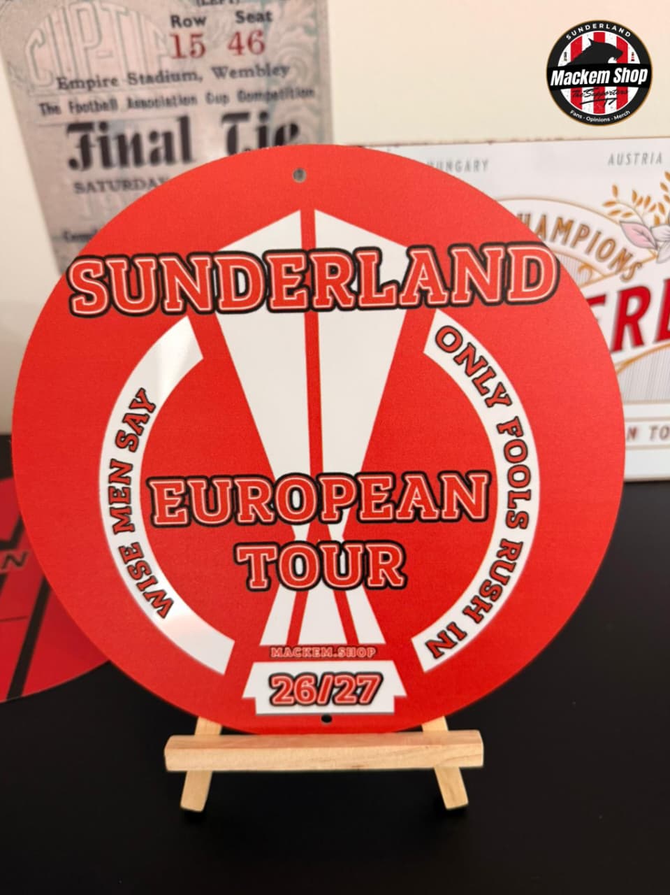 Red circular metal sign with 'Sunderland European Tour' text on a stand against a vintage background.