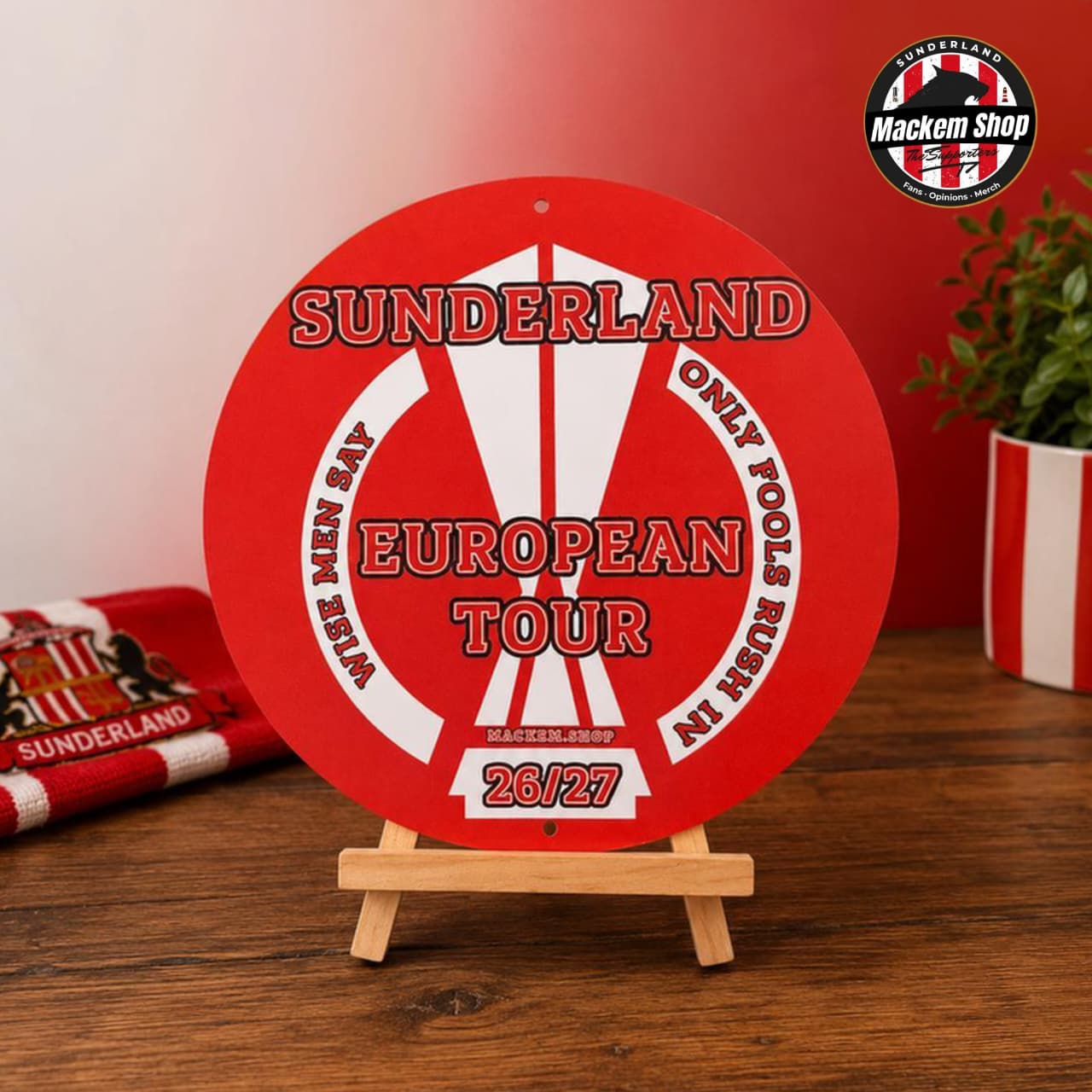 Red circular metal sign by MackemShop with 'Sunderland European Tour' text on a wooden stand against a red background.