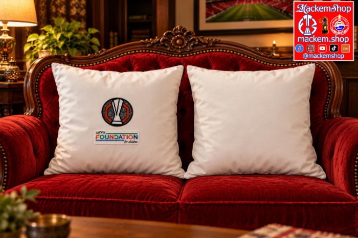 Two white square throw pillows with UEL Foundation patches, displayed on a red velvet sofa in a cozy, dimly lit living room setting, with MackemShop Logo visible on top right corner.