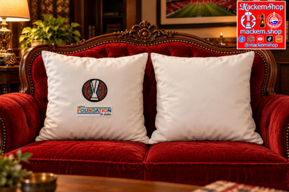 Two white square throw pillows with UEL Foundation patches, displayed on a red velvet sofa in a cozy, dimly lit living room setting, with MackemShop Logo visible on top right corner.