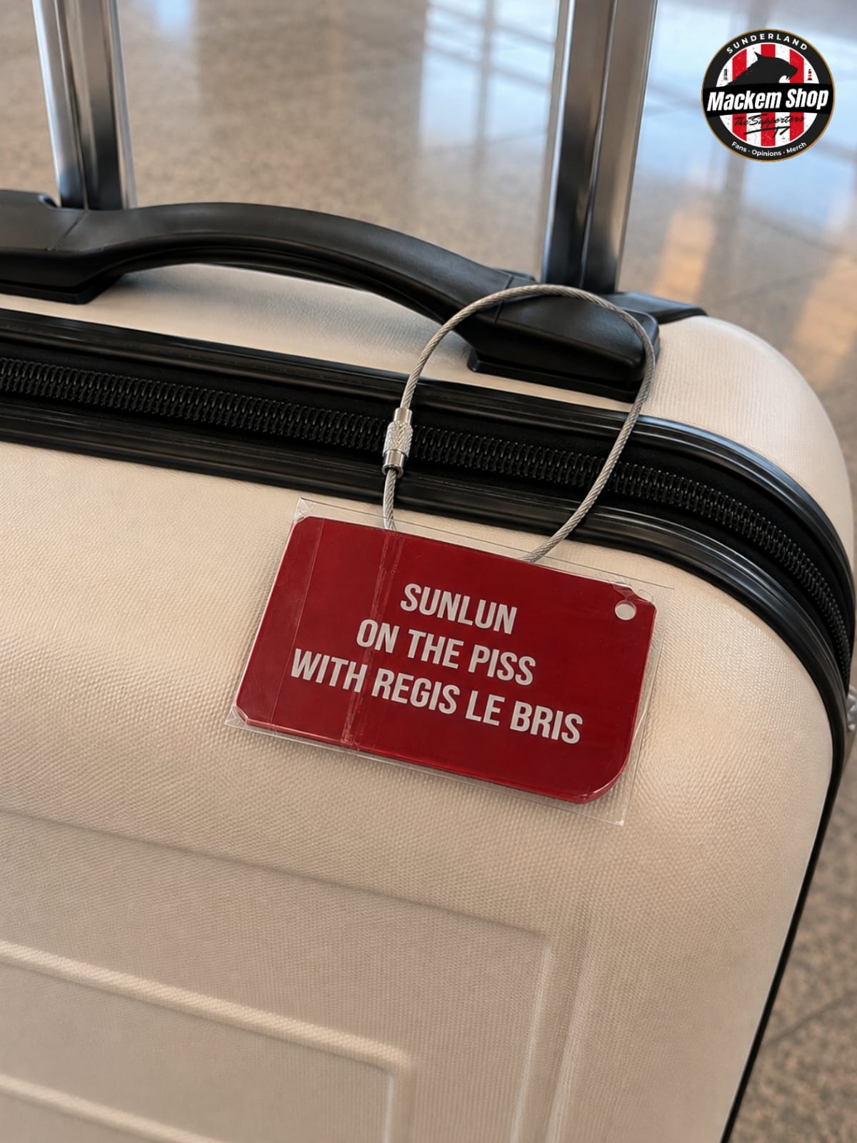 A close-up of a cream-colored hardshell suitcase with a black handle and zipper, featuring a red luggage tag that reads 'SUNLUN ON THE PISS WITH REGIS LE BRIS' in white text. A circular 'Mackem Shop' logo is visible in the top right corner.