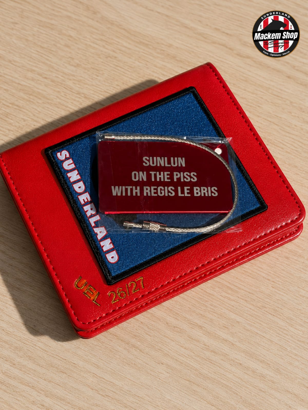 A red leather wallet or card holder on a light wood surface, featuring a blue insert with 'SUNDERLAND' text and a small red pouch labeled 'SUNLUN ON THE PISS WITH REGIS LE BRIS' containing a silver chain necklace. A circular 'Mackem Shop' logo appears in the top right.