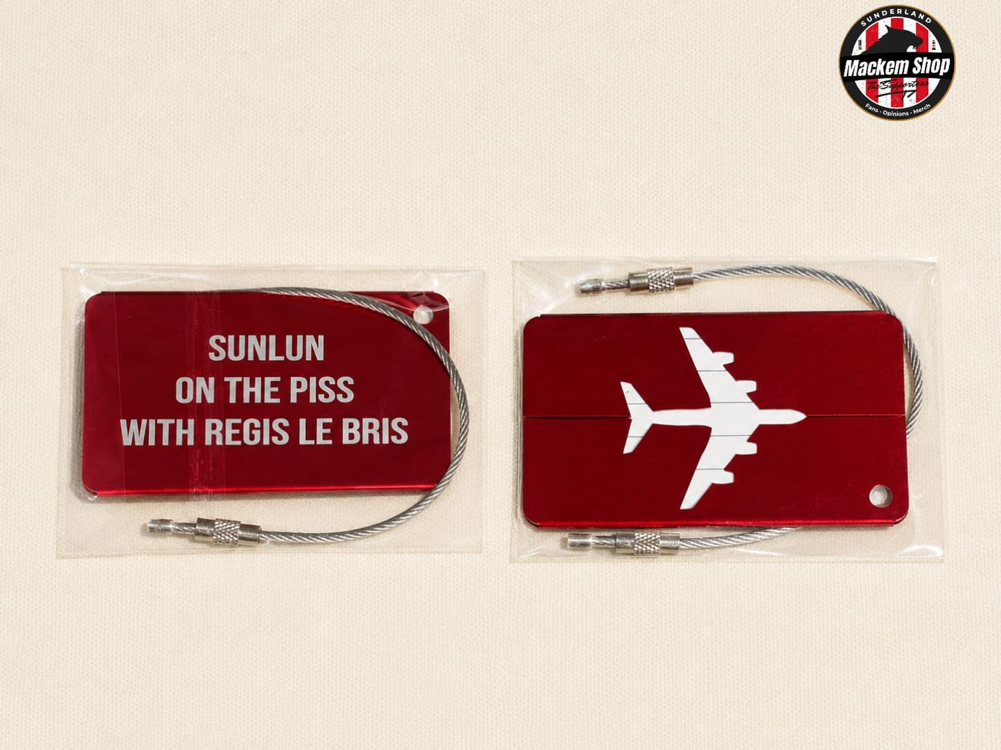 Two red metal luggage tags with silver cable locks, shown in clear plastic packaging on a light beige background. One tag has white text reading 'SUNLUN ON THE PISS WITH REGIS LE BRIS', and the other has a white airplane silhouette.