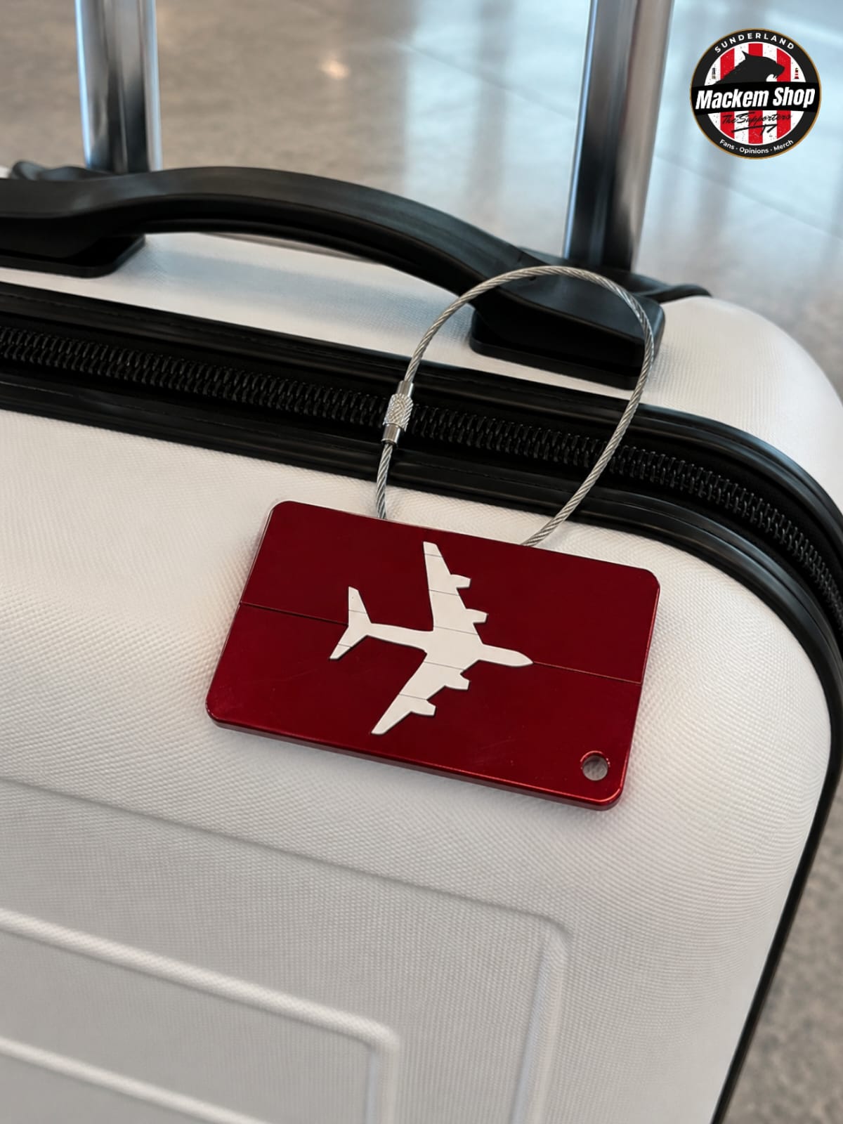 A rectangular red luggage tag with a white airplane silhouette, attached to a white suitcase handle by a silver wire loop. The tag has a small metal rivet and a circular cutout near the bottom right.
