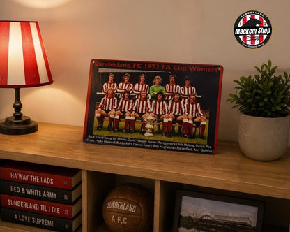 A rectangular Metal sign of the Sunderland FC 1973 FA Cup Winners team, displayed on a wooden shelf in a styled room setting with a lamp, potted plant, and books.