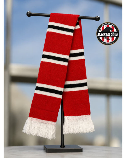 A red knit scarf with black and white stripes and white fringe tassels, displayed on a black stand. A circular 'Mackem Shop' logo is visible in the top right corner.